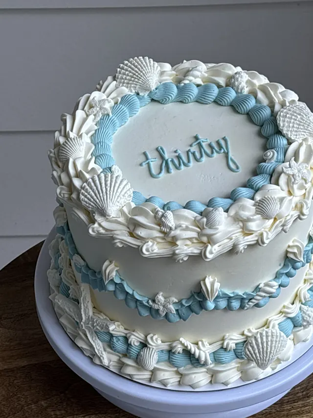 White round cake with blue piping, white seashells and "thirty."