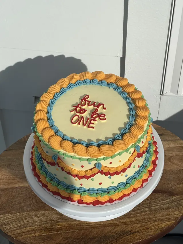 Round retro cake piped in orange, blue, green, red and yellow with "fun to be ONE."
