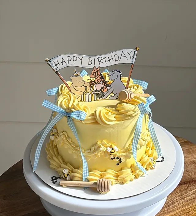 Yellow round cake with a Pooh "Happy Birthday" topper, gingham ribbons, bees and a honey dipper