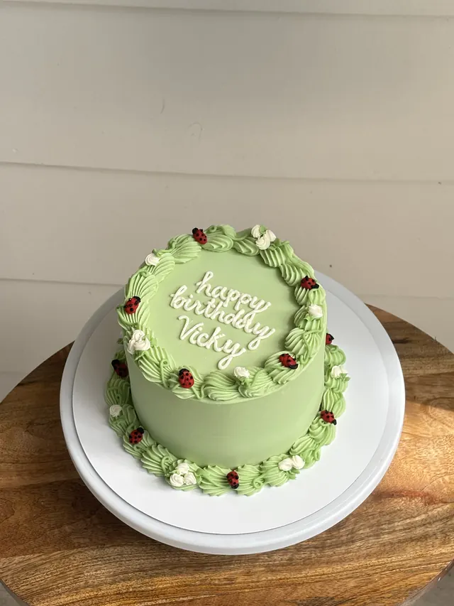 Sage green round cake with piped flowers, ladybugs and a birthday message