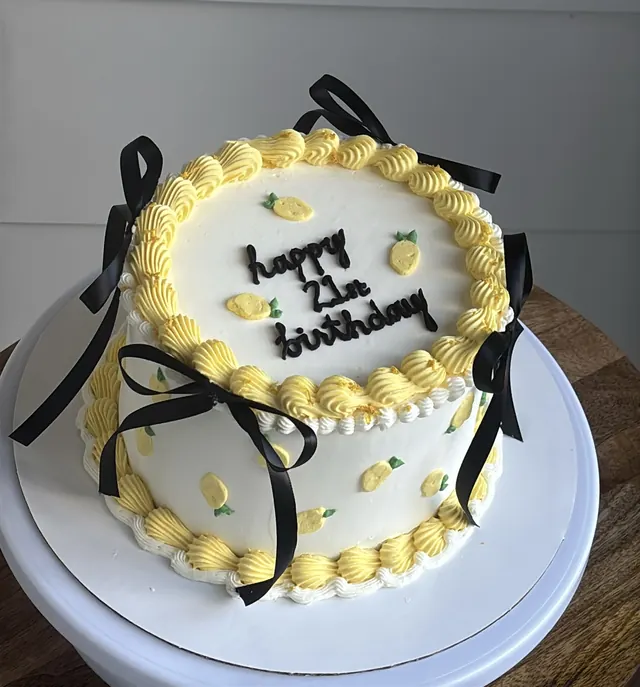 White round cake with yellow piping, piped lemons, black bows and "happy 21st birthday."