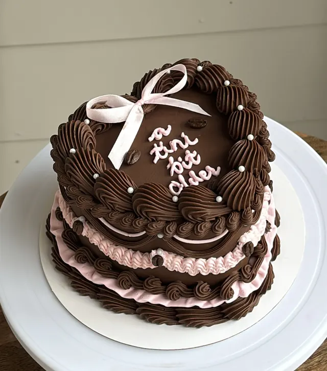 Chocolate heart cake with pink ruffle layers, a pink bow, coffee beans and "a tini bit older."