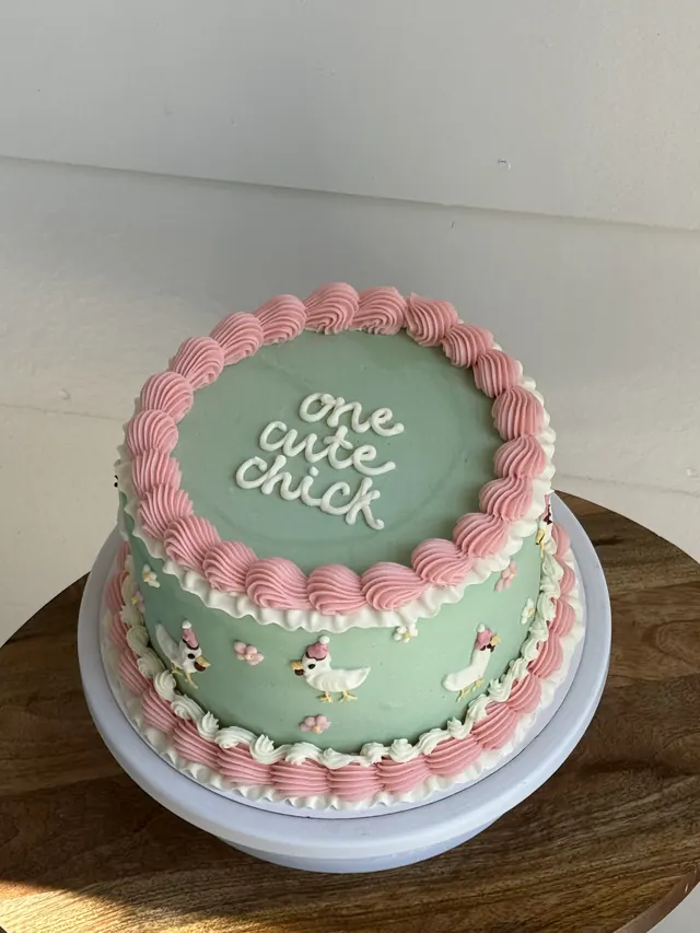 Sage green round cake with pink piping, piped chicks and "one cute chick."