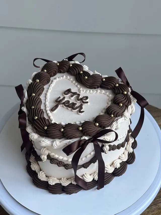 White heart cake with chocolate piping, brown bows, gold pearls and "one year."