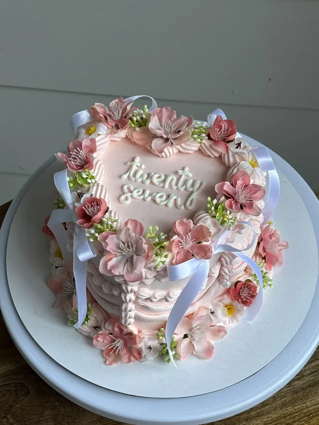 Pink heart cake with pink blossoms, white bows and "twenty seven."