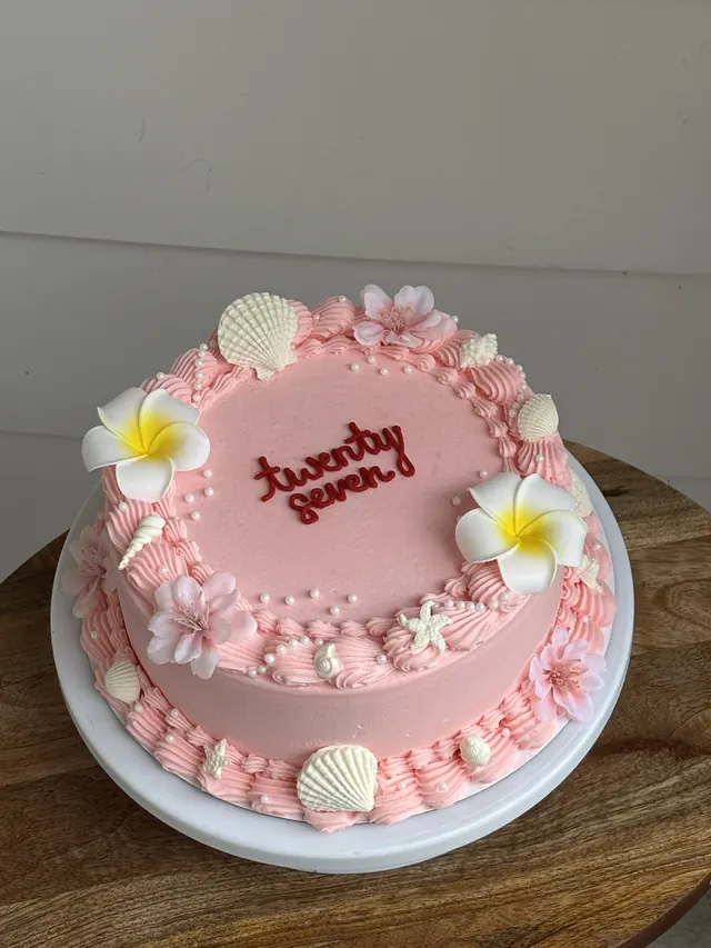 Pink round cake with plumeria flowers, seashells and "twenty seven."