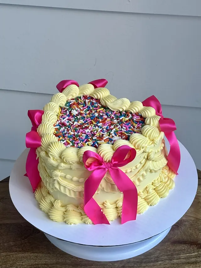 Pale yellow heart cake with a rainbow sprinkle top and hot pink bows