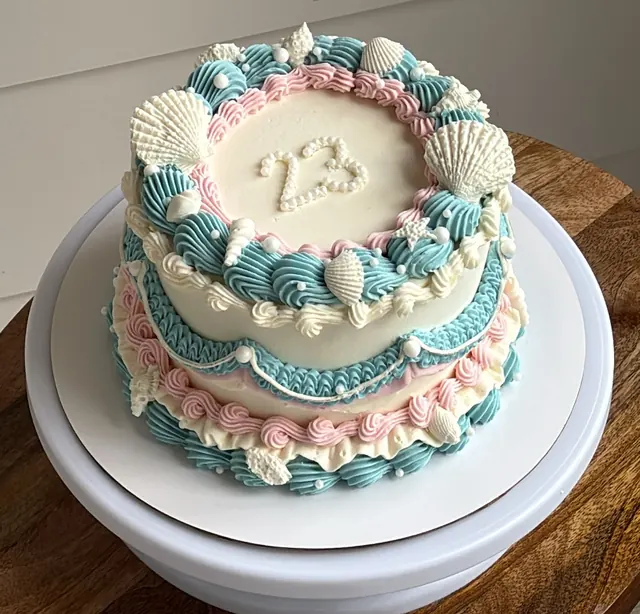 White round cake with teal and pink piping, white seashells and a 23