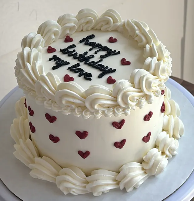 White round cake with ivory swirls, red piped hearts and "happy birthday my love."