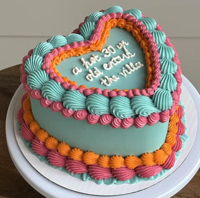 Teal heart cake with pink and orange piping and "a hot 30 yr old entered the villa."